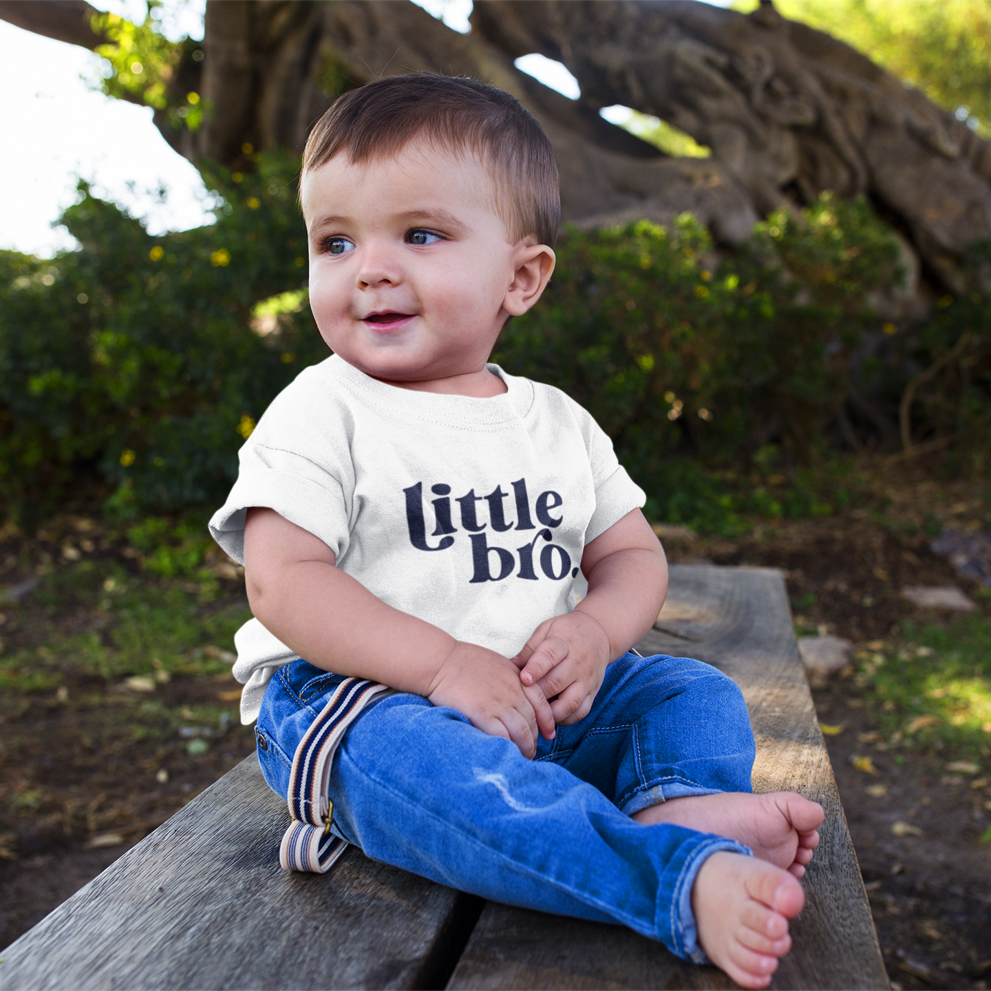 Child wearing a 'little bro' shirt sitting on a wooden bench outdoors.
