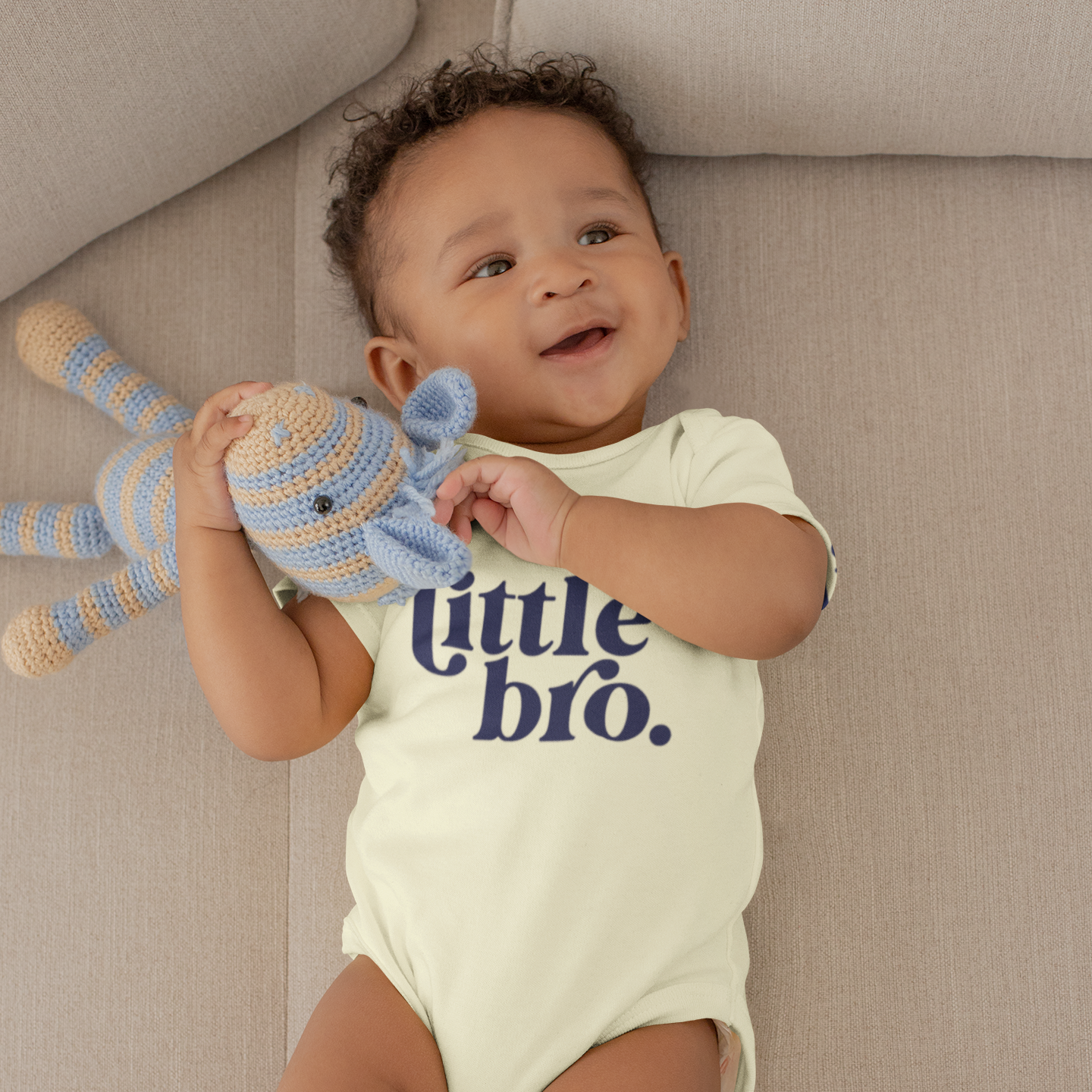 Baby wearing a 'little bro' onesie holding a toy on a couch